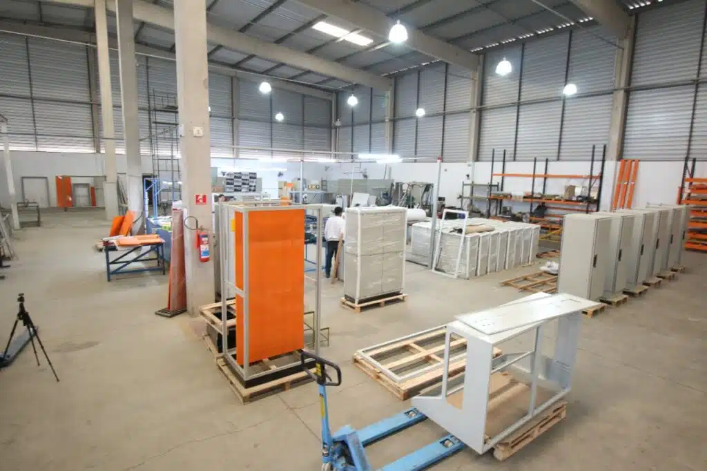 Interior do galpão de fabricação: invólucros de painel elétrico em paletes, estruturas metálicas em montagem, prateleiras de material e equipe trabalhando.